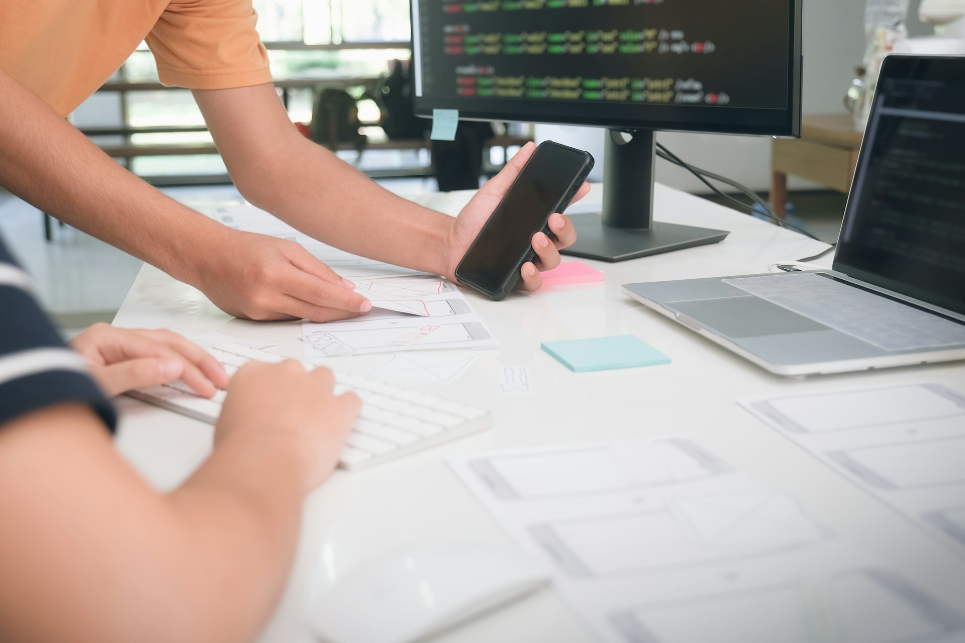 Two UX/UI designers collaborating on mobile app wireframes at a white desk, with smartphones, paper prototypes, and screens showing code and interface layouts