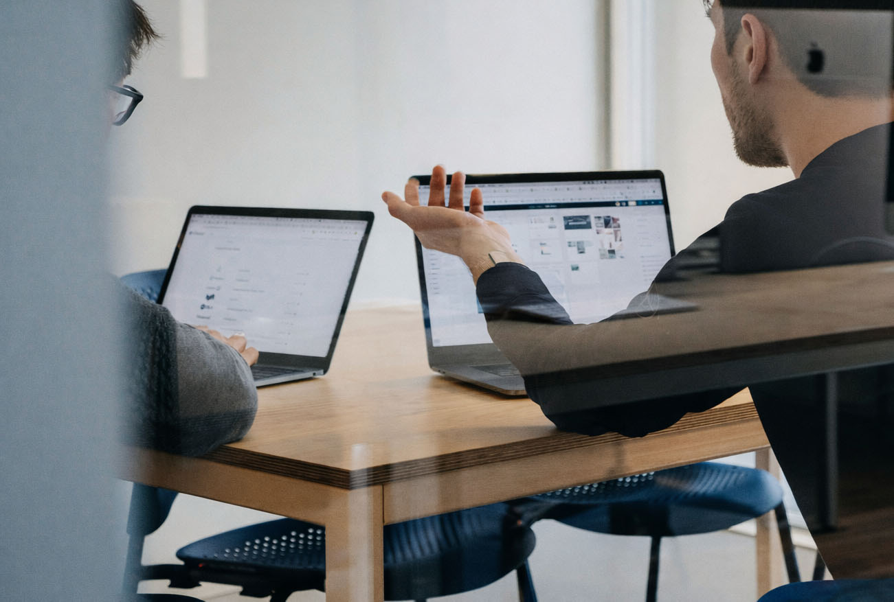 Two colleagues work at laptops in a bright office, seen through reflective glass.